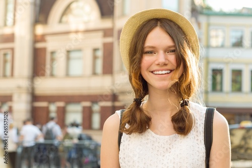 Summer outdoor portrait of smiling beautiful teenager girl 13, 14 years old wearing hat on city street, copy space