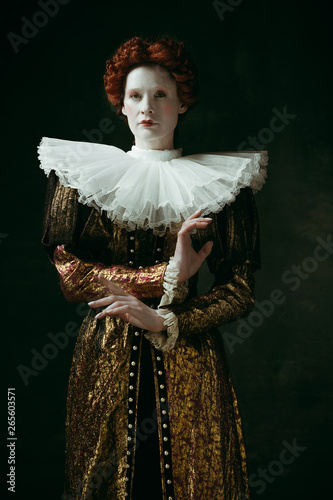 Flexibility of control. Medieval redhead young woman in golden vintage clothing as a duchess standing crossing hands on dark green background. Concept of comparison of eras, modernity and renaissance.