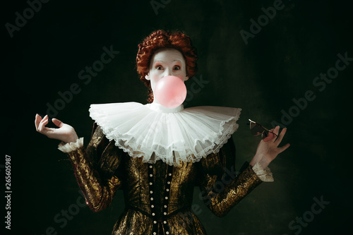 Medieval redhead young woman in golden vintage clothing as a duchess with red sunglasses blowing a bubblegum on dark green background. Concept of comparison of eras, modernity and renaissance.