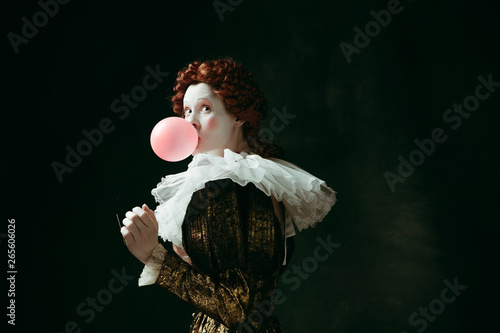 Medieval redhead young woman in golden vintage clothing as a duchess with red sunglasses blowing a bubblegum on dark green background. Concept of comparison of eras, modernity and renaissance.