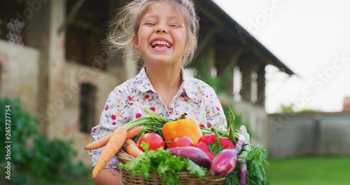 Portrait of happy little girl is holding a basket with fresh biologic just harvested vegetables and smiling in camera on a background of a countryside farm.