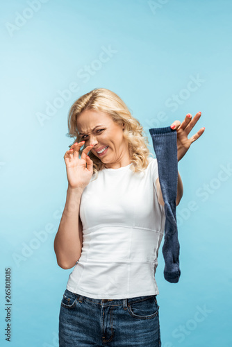 attractive curly blonde woman looking at smelly dirty sock on blue