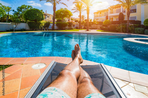 Relaxing at the swimming pool. Man relaxing next to swimming pool. Man enjoying the hot summer at swimming-pool. Sunbathing by the swimming pool, mans legs lying down on a sun lounger over the water.