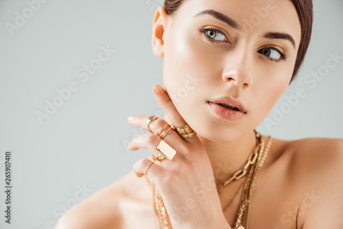 young nude woman in golden rings and necklaces looking away isolated on grey