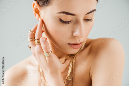 young naked woman with shiny makeup in golden necklaces and rings isolated on grey