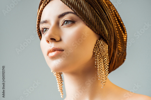 young naked woman with shiny makeup, golden rings in turban looking away isolated on grey