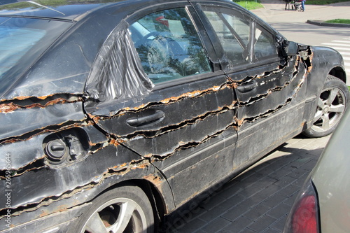 An old, rusty, thrice cut along the long side, a black car drives through the city streets. Sunny day.