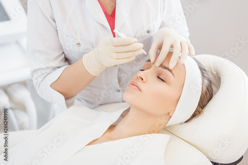 Unidentified master cosmetologist makes botox injections into the eyebrow of a beautiful young woman client. The concept of rejuvenating aesthetic procedures