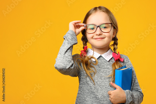 Portrait of a cute little kid girl on a yellow background. Child schoolgirl looking at the camera, holding a book and straightens glasses. The concept of education. Copy space.