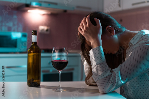 Unhappy, lonely, young woman with red wine is drinking alone at home in evening. Life difficulties and problems. Female alcoholism and alcohol addiction