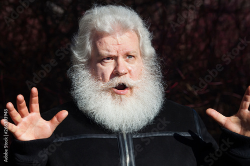 Amazed senior man making a helpless gesture against dark natural background. Close up sunny portrait of elderly stylish male with splendid mustache and beard wearing black flight (bomber) jacket.