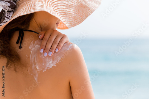 Young girl in straw hat is applying sunscreen on her back to protect her skin