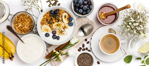 Oat granola with yogurt, honey, fresh bananas, blueberries, chia seeds in bowl and cup of coffee on white background