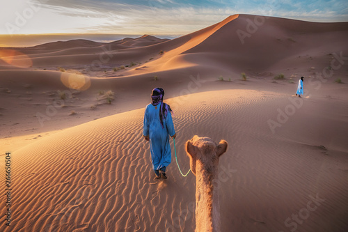 Two Tuareg nomads dressed in traditional long blue robes, lead a camel through the dunes of the Sahara Desert at sunrise in Morocco.