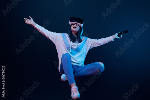 happy woman in virtual reality headset levitating and holding joystick on blue