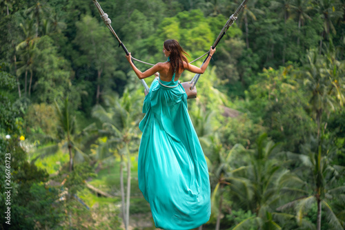 Tanned young woman riding on a long swing. Island of Bali. Tropical forest on the background. Travel and joy. Close up