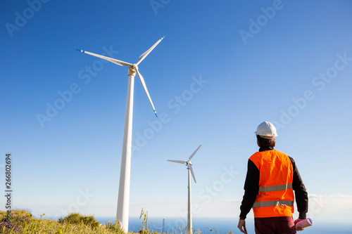 Ingegnere elettrico controlla l'impianto eolico con il gilet ad alta visibilità e casco bianco. Progetto in mano. Concetto di manutenzione