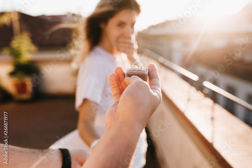 Man holding wedding ring in front of astonished happy girl covering mouth with hand. Romantic photo of charming woman standing on roof early in evening on date with boyfriend in anniversary.