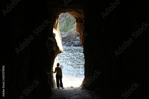 a young woman stands in a dark cave and watches at a blue sea outside