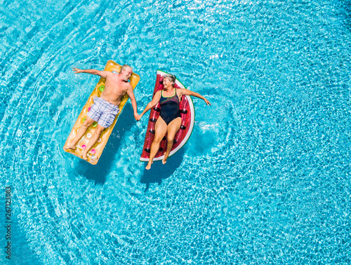 Above vertical view of people old senior couple taking hands with love and having fun on the blue clear swimming pool together enjoying the summer holiday vacation with trendy coloured lilos mattress