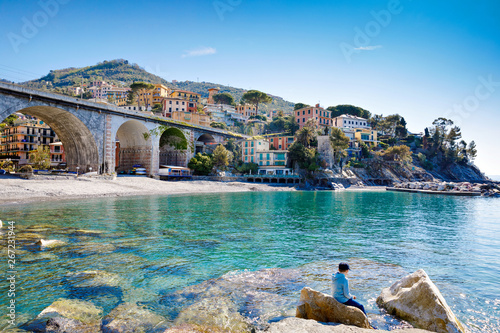 Little kid boy climbing on stones on beach of Mediterranean sea in Liguria region, Italy. Awesome landscape of Zoagli, Cinque Terre and Portofino. Beautiful Italian city with colorful houses.