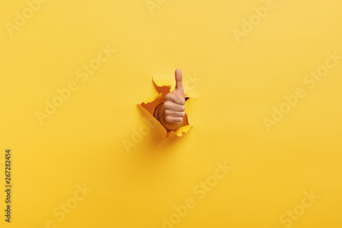 Image of unrecognizable man makes thumb up gesture, demonstrates approval or agreement, gestures through torn paper wall yellow background. Body language concept. Hand sign. Hole in wall. Like gesture