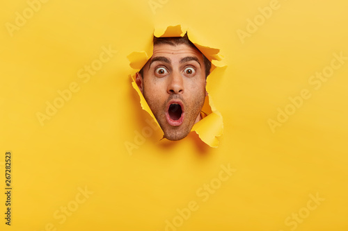 Stupefied young man with bristle keeps jaw dropped from surprisement, has popped eyes, stares through yellow torn paper, impressed by shocking rumors. People, human emotions and reaction concept