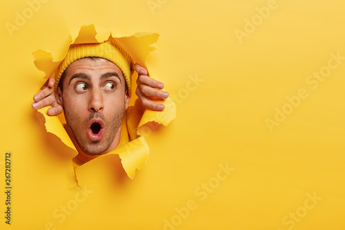 Surprised male face through paper hole. Emotional astonished young man wears yellow headgear, makes slot in background with hands, keeps mouth wide opened. Copy space to insert yout text or slogan