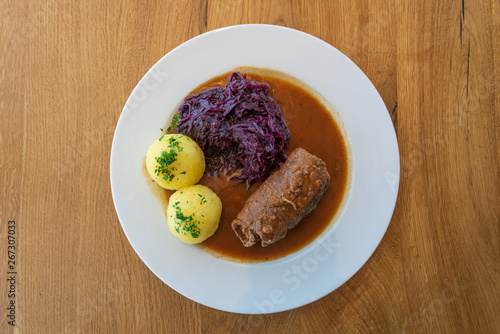 Rinderroulade, Beef roll or roulades, traditional german meal, thin meat wrap bacon, onion and pickle, serve with pickled red cabbage, dumplings potatoes and sauce on white plate and wooden table.