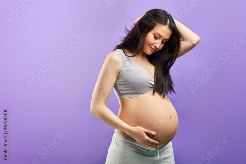 Pregnant woman in third trimester, dressed in comfotable home wear, posing against studio purple wall, looks happily at camera, being in anticipation of baby. Happy pregnancy and motherhood concept