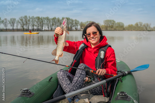 Kayak fishing. Fisher girl holding pike fish trophy on inflatable boat with fishing tackle at lake.