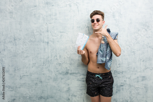 Young man wearing a swimsuit holding air tickets showing a mobile phone call gesture with fingers.