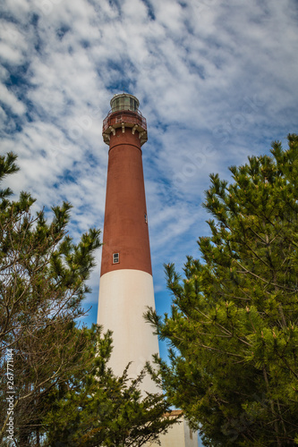Barnegat Lighthouse on Long Beach Island, NJ, surrounded by large evergreen pine trees on a sunny spring day with blue sky dotted with clouds