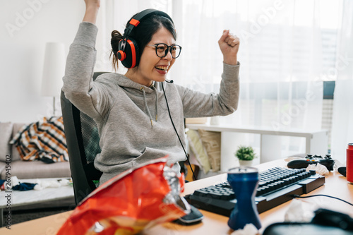 Professional gamer girl putting on headset and starts playing online video game on personal computer. Casual asian geek woman. happy female in eyeglasses winning pc game at home with yes hand gesture