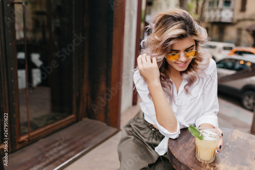 Portrait of relaxed young woman in white shirt and beige skirt plays with her fair hair and enjoys cold cappuccino. Amazing blonde girl chilling in outdoor cafe and drink tea with mint and ice cubes.