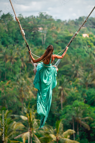 Opalona młoda kobieta jedzie na długiej huśtawce w długiej turkusowej sukni. Wyspa bali. Las tropikalny w tle. Podróż i radość