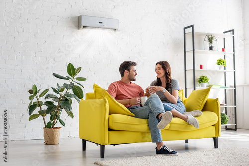 happy young couple talking and holding cups while sitting on yellow sofa under air conditioner at home