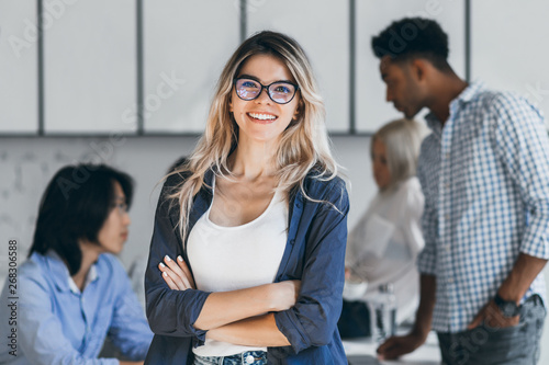 Confident blonde female manager posing with smile after conference with other employees. Asian programmer talking with african freelancer while fair-haired secretary laughing on foreground.