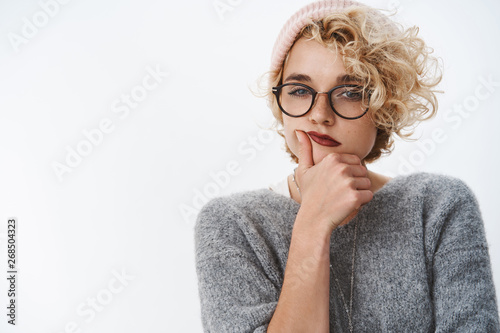 Indoor shot of picky and thoughtful creative female designer making up new ideas wearing warm beanie and winter sweater with glasses squinting as thinking holding hand on chin over white background