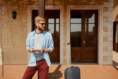 Picture of unshaven attractive young man with thick beard waiting for check in outside guest house with suitcase, killing time, playing video games and surfing social networks on digital tablet