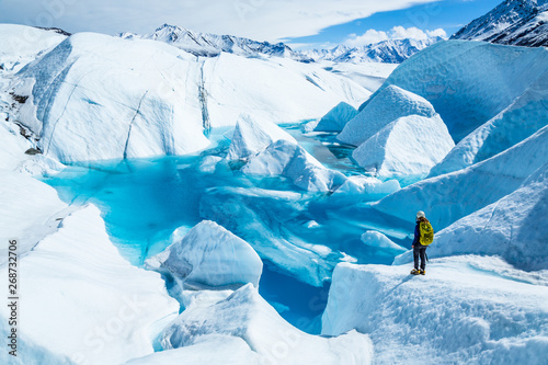 Young woman standing near deep blue lake on the Matanuska Glacier in Alaska. She wears a backpack and helmet with ice axe in hand for summer glacier travel.