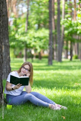 Young blonde woman with glasses sit under the tree in summer park and read book with sceptic face. Cap of coffee near. Student lifestyle. Space for text