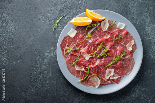 Marbled beef carpaccio with arugula, lemon and parmesan cheese on dark concrete table. Top view, flat lay with copy space