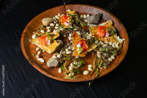Slices of salmon on scraps of cakes with spices are laid out on a wooden dish decorated with peanuts, stones and sprigs.