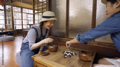 Two beautiful girl friends playing chess with japanese style house interior. young female travelers having fun together with igo go stones. attractive laughing women play chinese board game cheerful