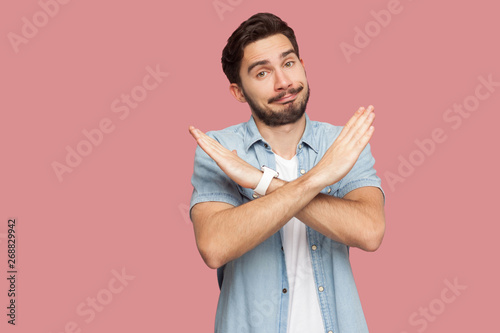 Portrait of serious handsome bearded young man in blue casual style shirt standing with X sign hands and looking at camera. indoor studio shot, isolated on pink background.