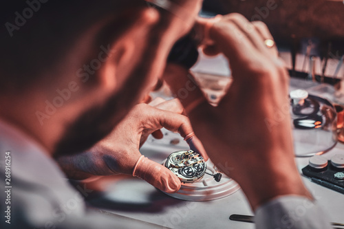 Mature clockmaster is fixing old watch for a customer at his busy repairing workshop.