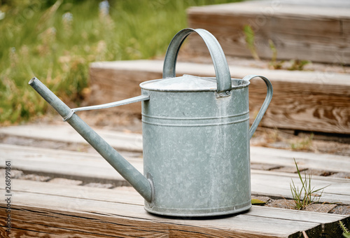 Old metal watering can standing on the wooden stairs of a terrace in the garden with grass and plants in the background