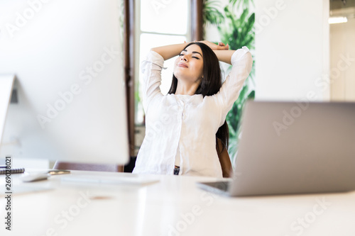Cheerful pretty woman sitting at desk with hands behind head. Business lady imagines happy future with closed eyes. Relaxed girl resting in office chair. Great pleasure from business success concept