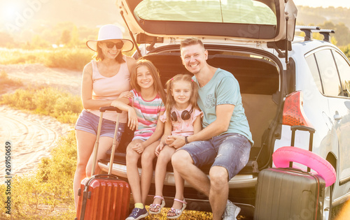 Happy pleasured family sitting on a trunk of a car with suitcases on a travel vacation